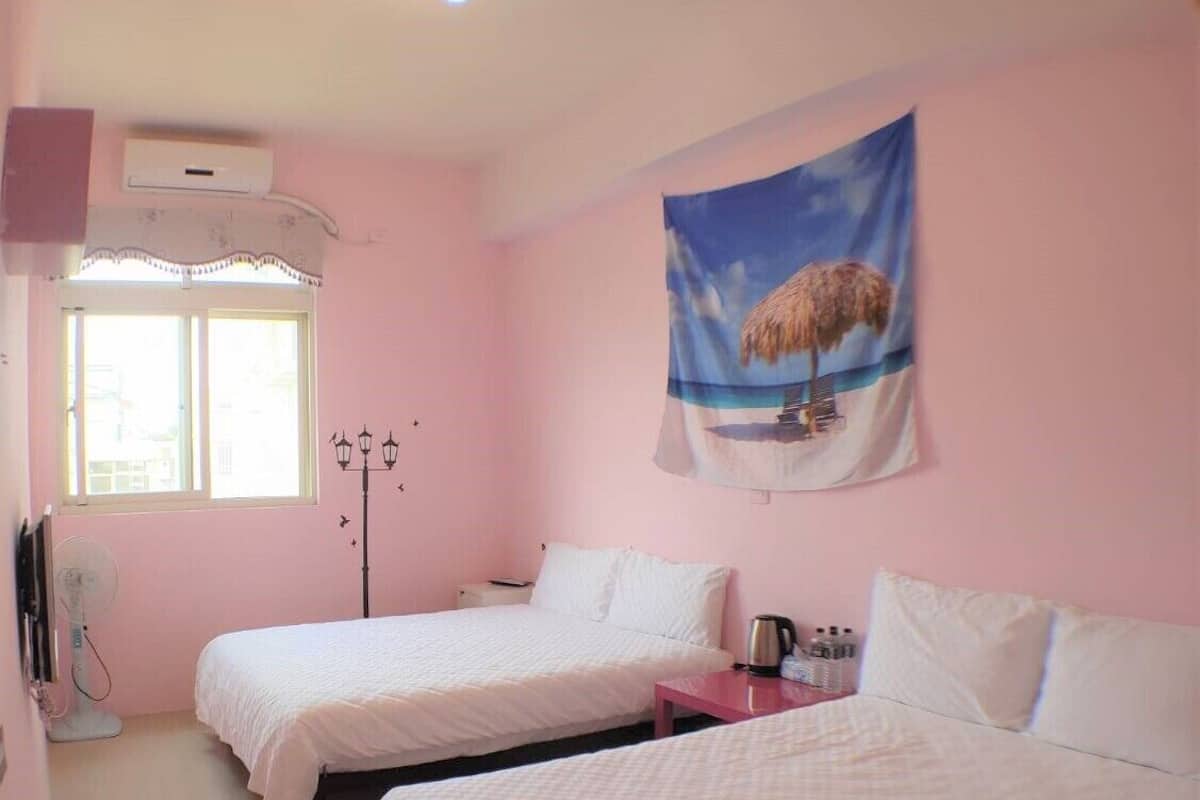 A room with two beds, a window, an air conditioning unit, a wall-mounted lamp, and a beach-themed tapestry.