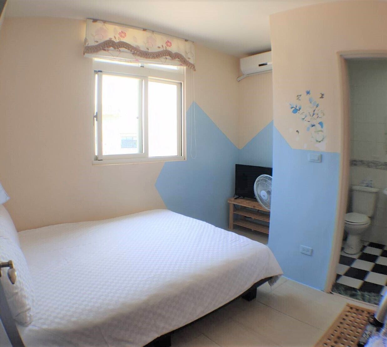 A bedroom with a bed, a window, a fan, and a bathroom with a toilet and sink.