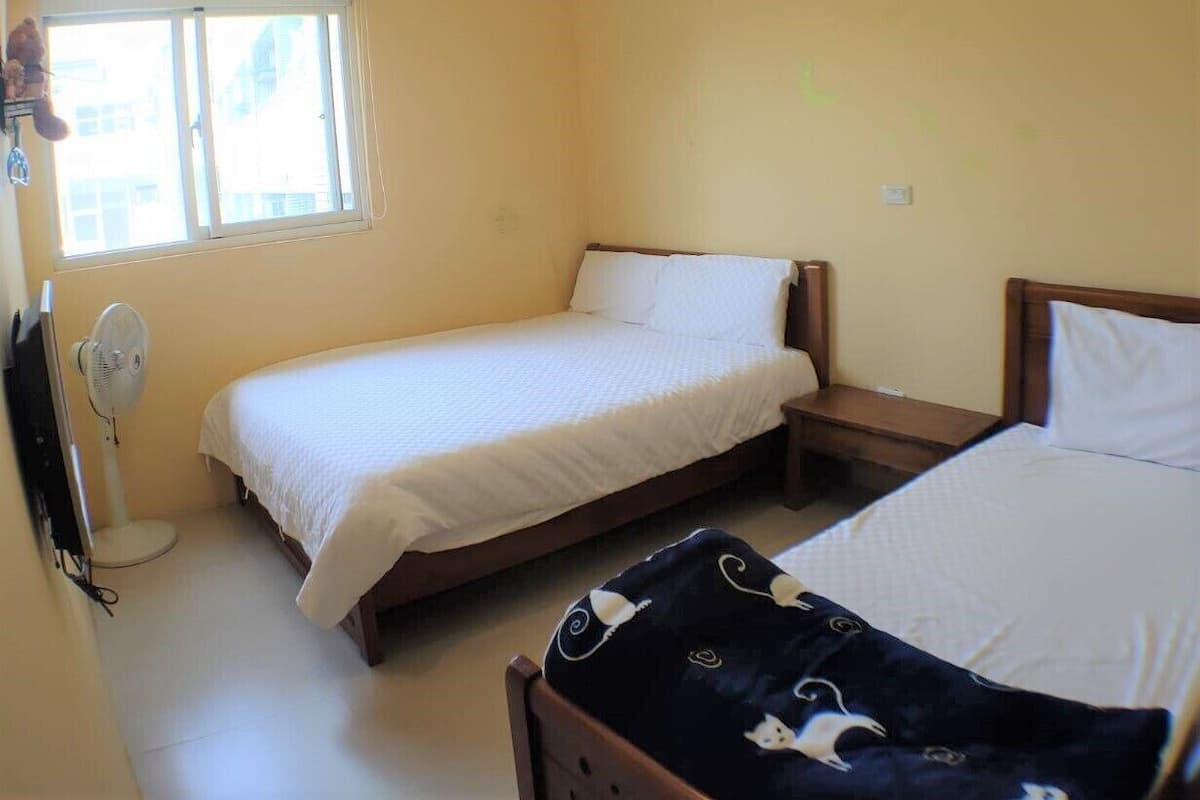 A room with two single beds, a standing fan, a window with a curtain, and an air conditioning unit.