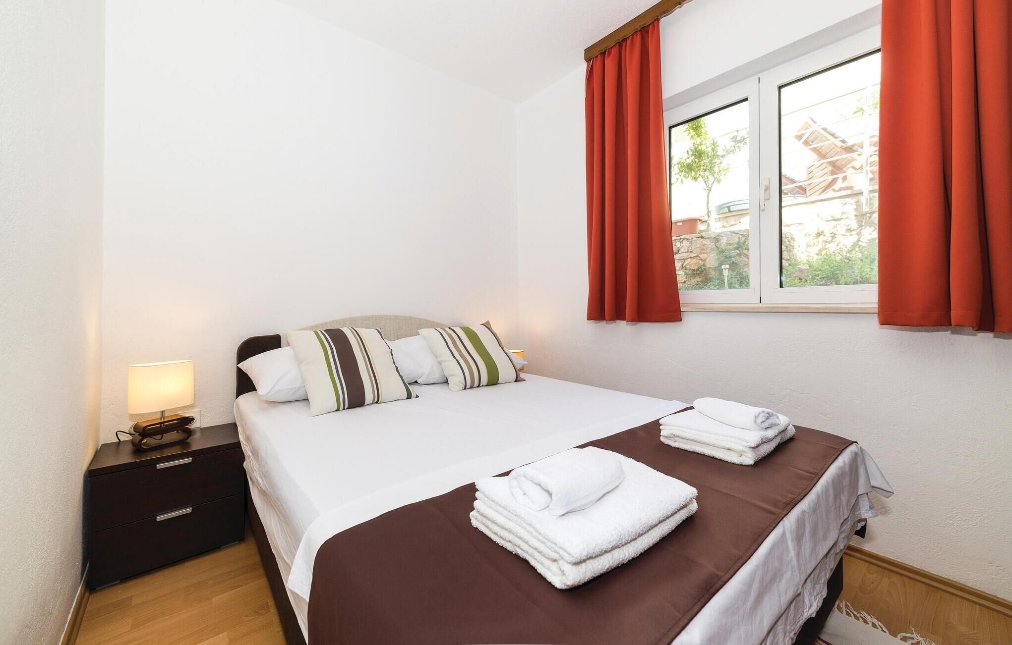 A hotel room with a bed, a nightstand, a lamp, a window with curtains, and a view of buildings outside.
