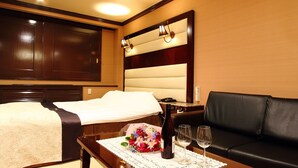 Una habitación de hotel con una cama, un sofá de cuero negro, una mesita con un arreglo floral y dos copas de vino, y una lámpara fijada a la pared.