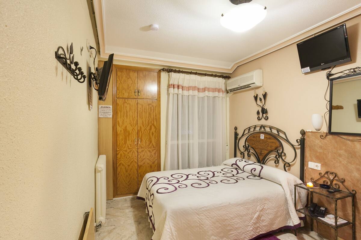 A bedroom with a bed, wooden wardrobe, air conditioning unit, and a television on the wall.