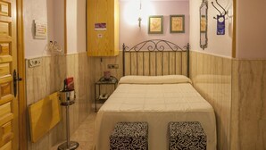 A bedroom with a bed, two patterned ottomans, a wooden wardrobe, a small table with a telephone, and a wall-mounted radiator. - Pensión San José (Bullas)
