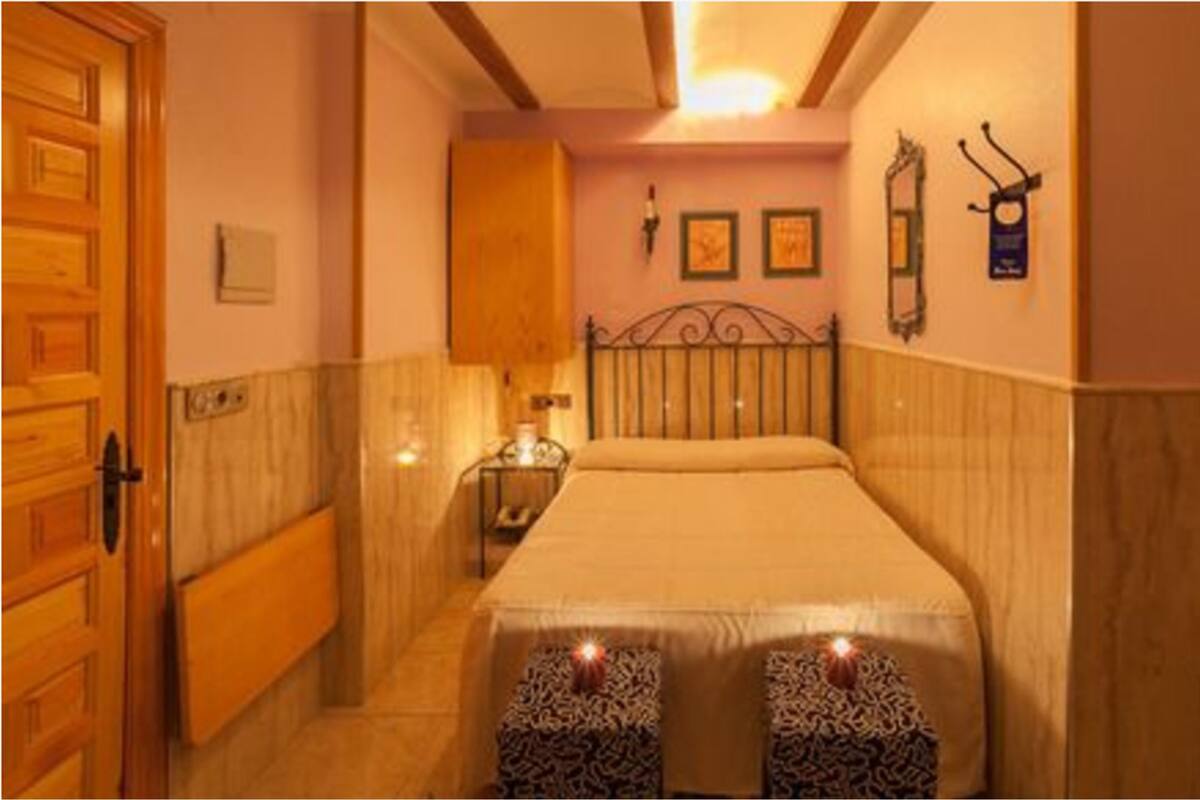 A bedroom with a bed, wooden wardrobe, bedside table, wall-mounted lamp, and framed pictures.