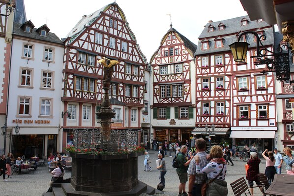 Una plaza tradicional europea con edificios de entramado de madera, una fuente central y zonas de estar al aire libre.