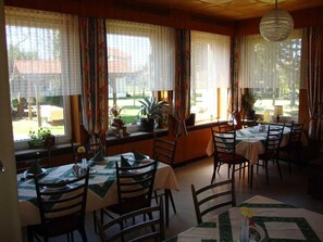 A dining area with tables set for four, large windows with curtains, and a view of the outdoors.