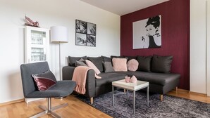 Ein modernes Wohnzimmer mit einem grauen Sofa, einem schwarzen Sessel, einem kleinen weißen Tisch und einem gemusterten Teppich. An den Wänden befinden sich Kunstwerke und eine Wand in Rot gehalten.