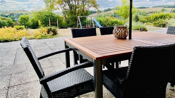 Eine Terrasse mit Tisch, Stühlen und Sonnenschirm, mit Blick auf einen Spielplatz und Hügel in der Ferne.