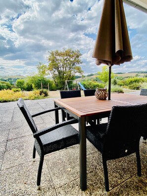 A patio with a table and chairs, an umbrella, and a view of a playground and hills in the distance. - Villa Schoenau Apartment 1 with stunning views, terrace and large garden (Bad Münstereifel)