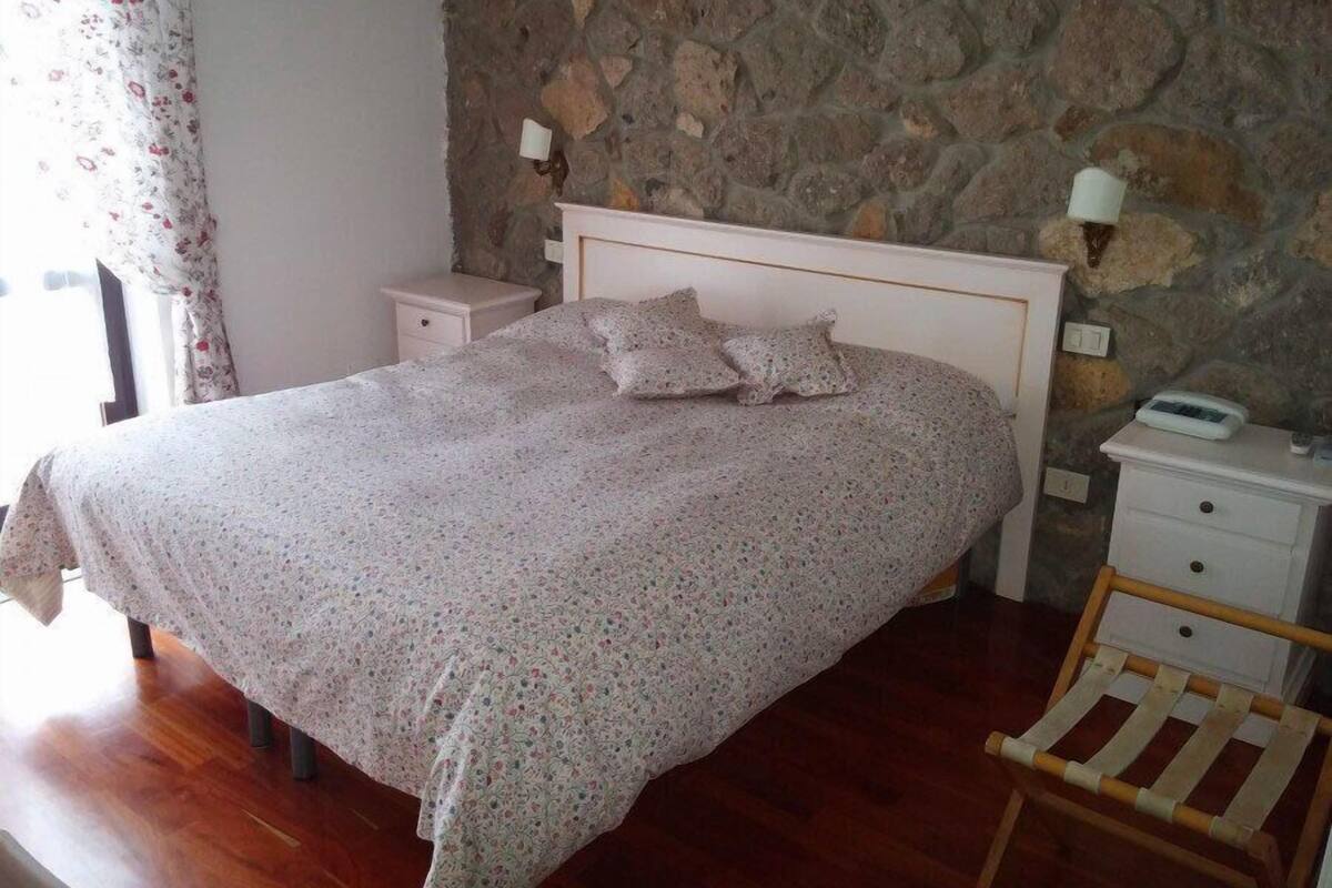 A bedroom with a stone wall, a wooden bed frame, a nightstand with a lamp, and a chair.