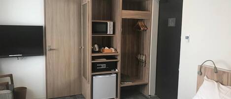 A hotel room with a bed, a TV mounted on the wall, a wardrobe with a microwave, a small fridge, and a coat rack.