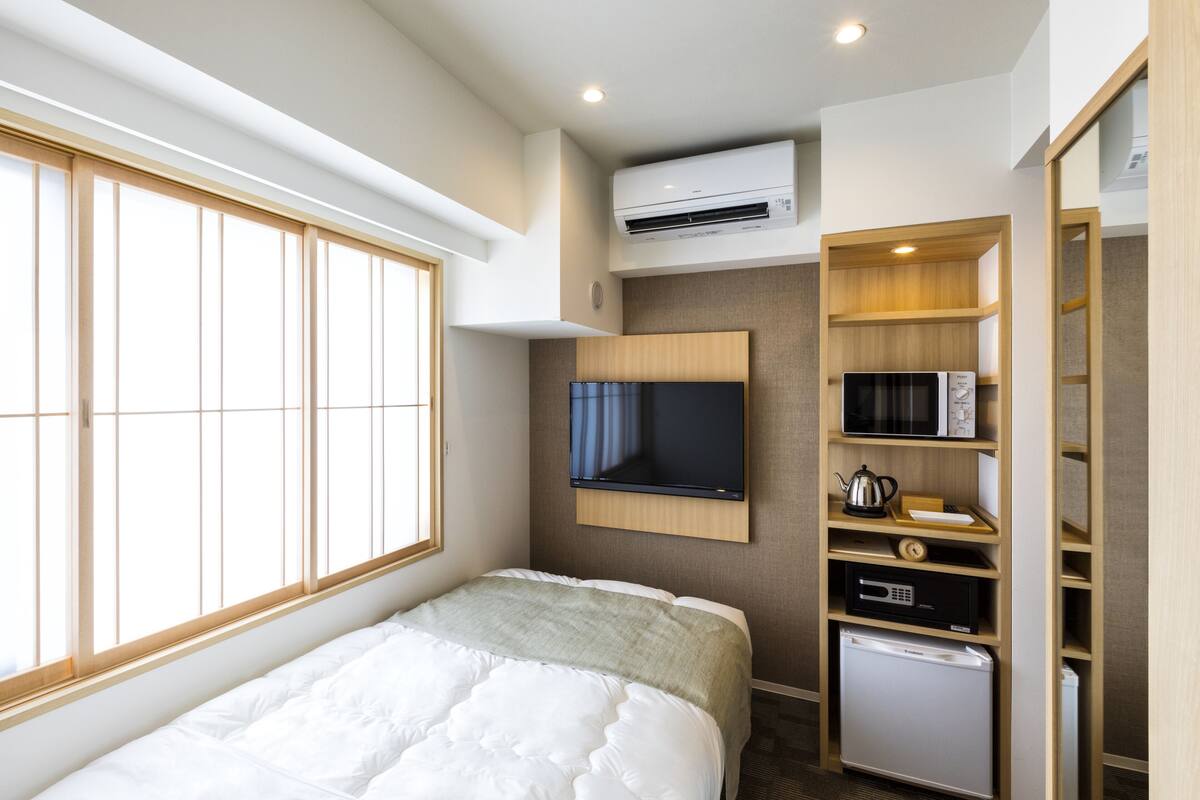 A hotel room with a bed, a television, an air conditioning unit, a microwave, a refrigerator, and a wooden cabinet.
