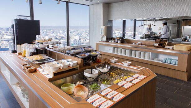 A modern breakfast buffet with a city view, featuring various food items and utensils. - New Tomakomai Prince Hotel Nagomi (Tomakomai)