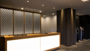 A reception area with a modern design, featuring a wooden reception desk with a bright illuminated panel, a decorative partition with geometric patterns, and a tall vertical panel with vertical lines. - New Tomakomai Prince Hotel Nagomi (Tomakomai)