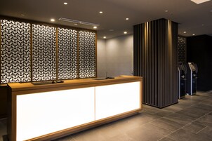 Un área de recepción de diseño moderno, con un mostrador de madera y un panel iluminado brillante, una partición decorativa con patrones geométricos y un panel vertical alto con líneas verticales.