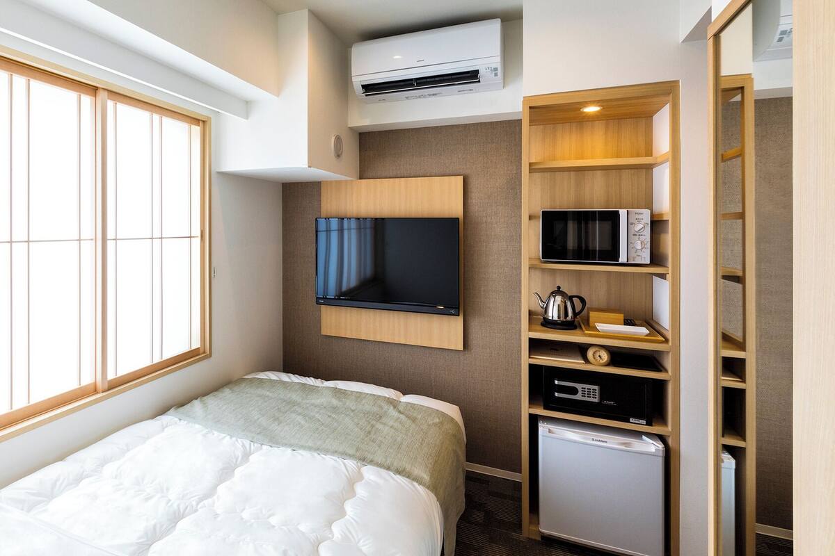A hotel room with a bed, a TV mounted on the wall, a microwave, a refrigerator, and a window with blinds.