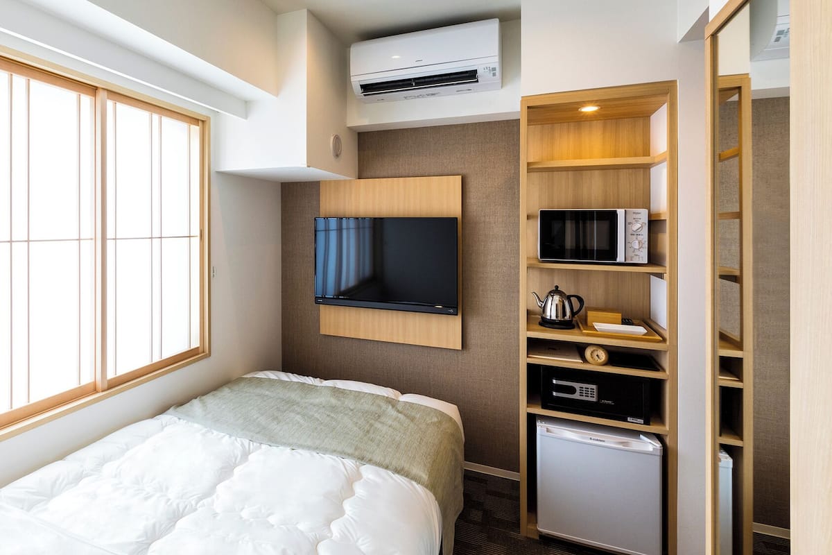 Quarto de hotel com cama, televisão fixada na parede, micro-ondas, frigorífico e janela com persianas.