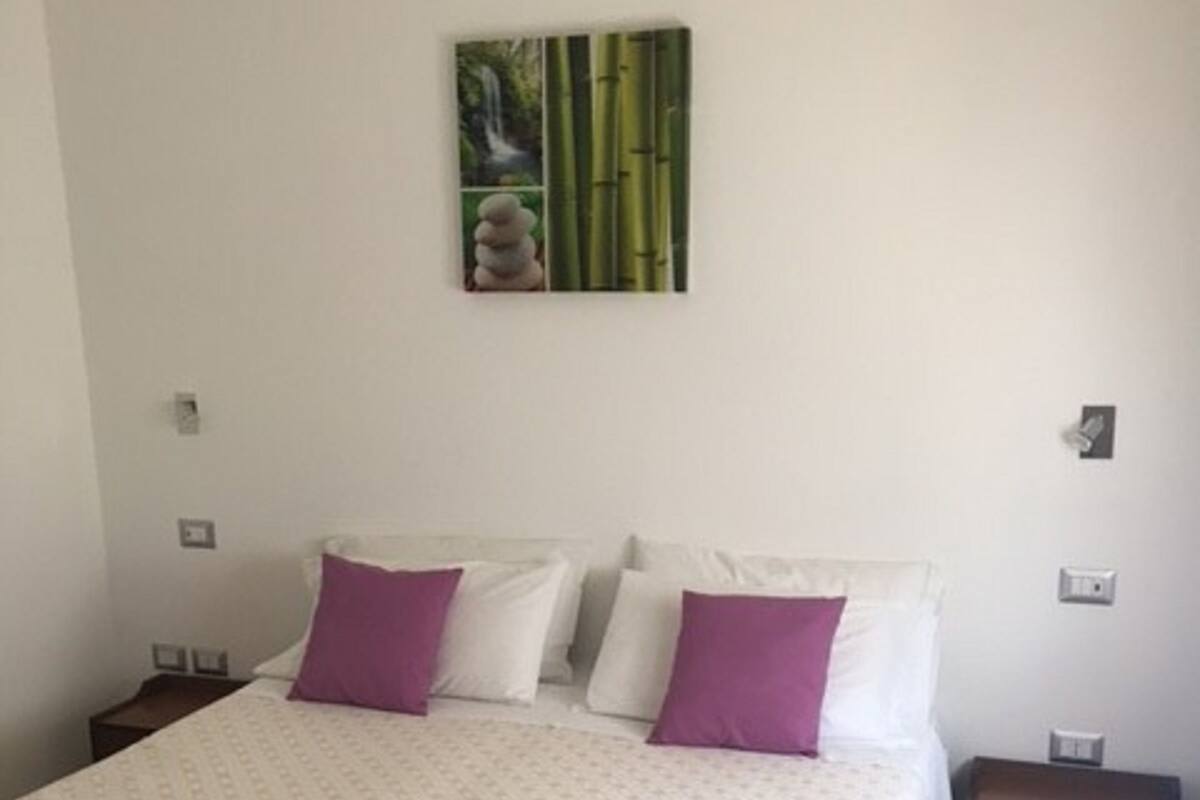 A bedroom with a bed, two nightstands, a wooden chest, and a wall-mounted artwork.