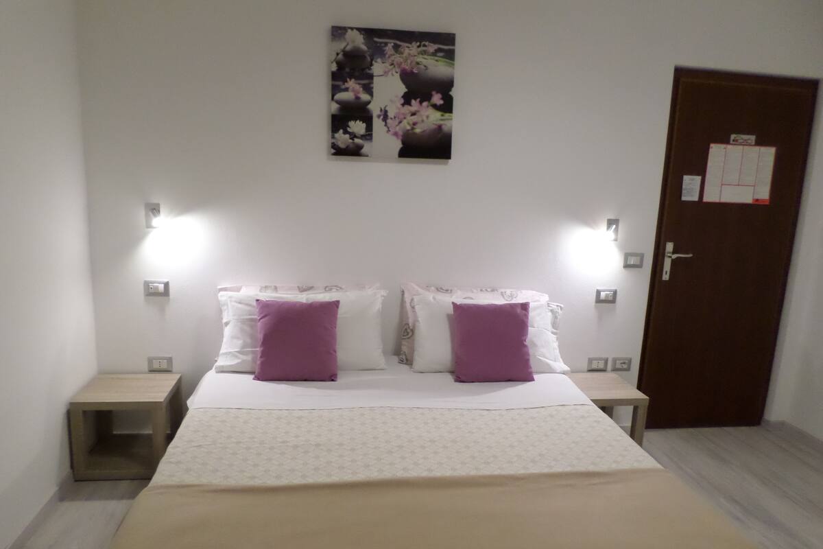 A neatly made bed with a patterned bedspread and two purple pillows, flanked by two bedside tables with lamps.