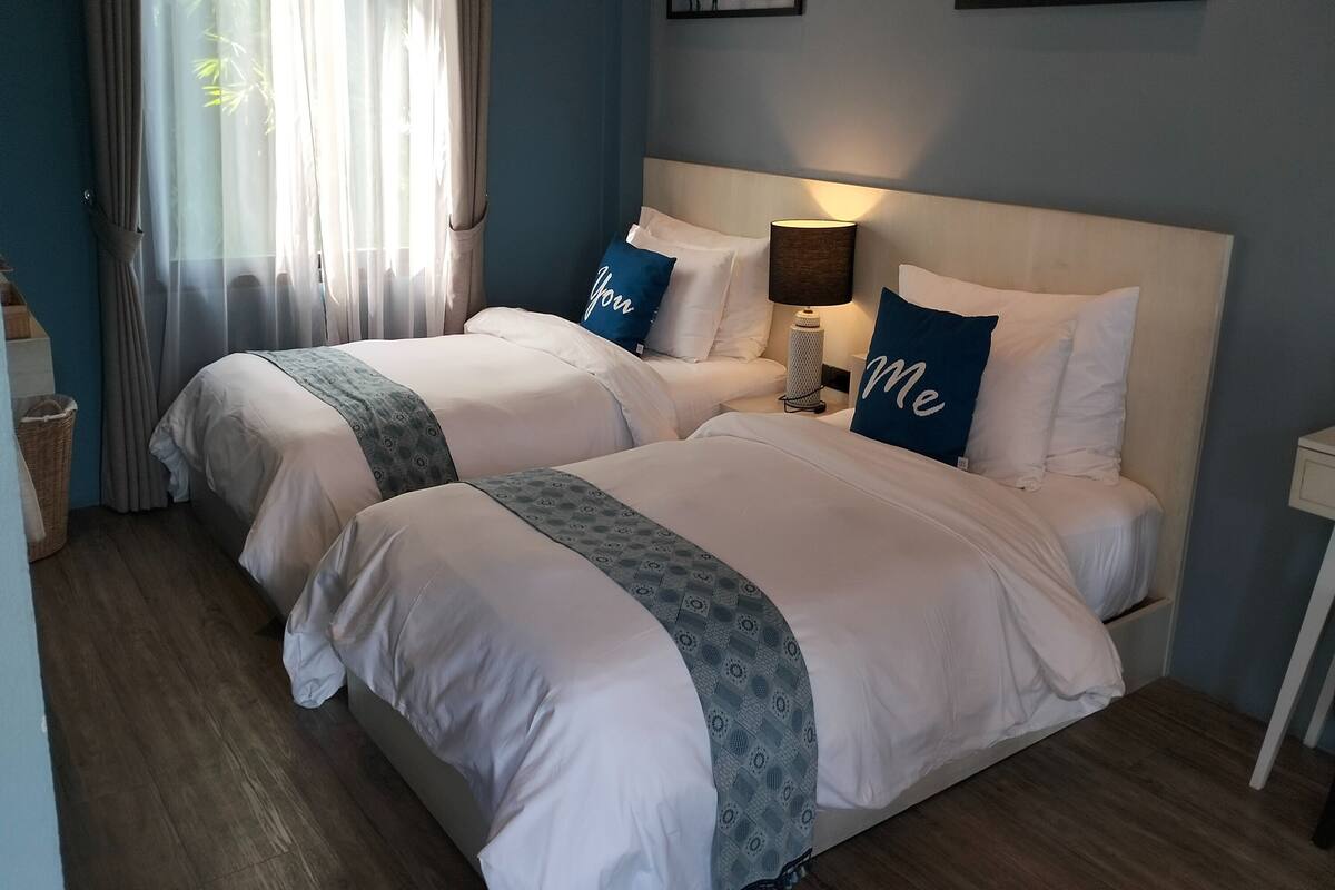 A hotel room with two beds, each with white bedding and blue pillows. There is a lamp on the nightstand, a basket on the floor, and a window with sheer curtains.