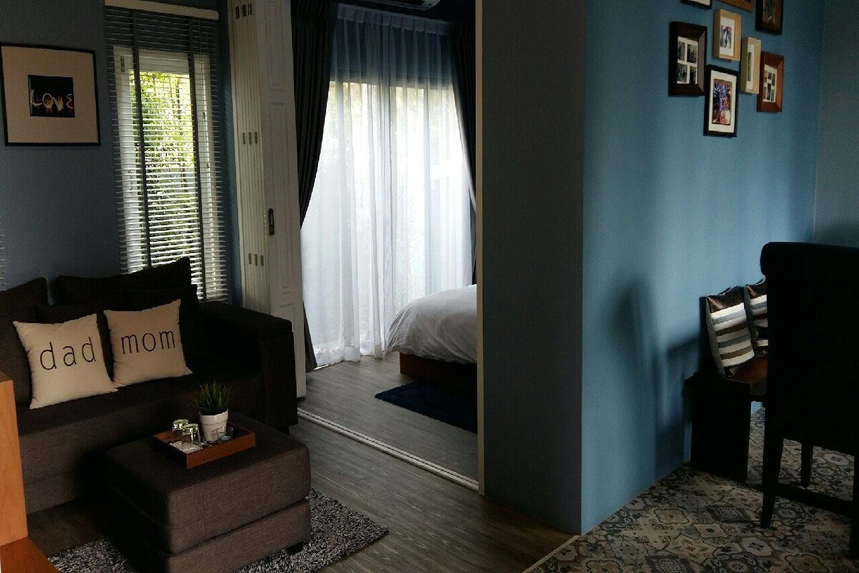 Una sala de estar moderna con un sofá, un reposapiés y una pequeña mesita de noche con una planta. Se ve un dormitorio con una cama y cortinas a través de una puerta abierta.