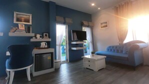 Una sala de estar con un sofá azul, una mesa de centro blanca, un televisor sobre un mueble, un refrigerador pequeño y una silla.