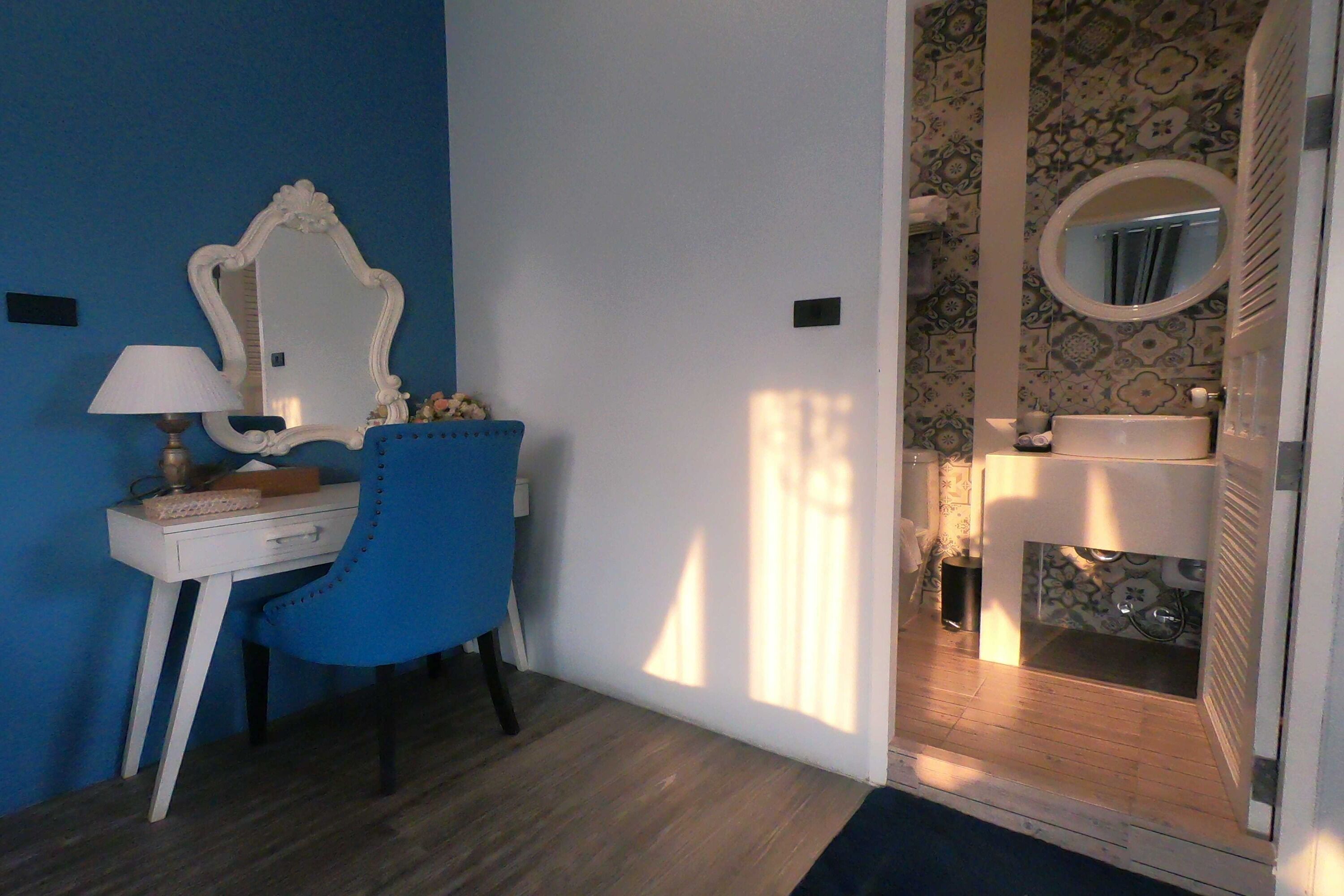 A room with a blue wall, a white desk with a lamp, a blue chair, and a bathroom with a sink and mirror.