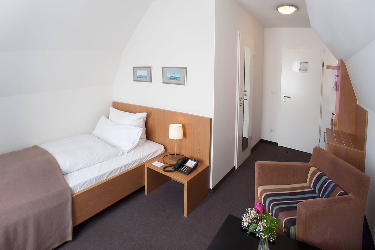 Une chambre d’hôtel comprenant un lit, une table de chevet avec une lampe, une chaise, une petite table avec des fleurs et une porte donnant sur une autre pièce.