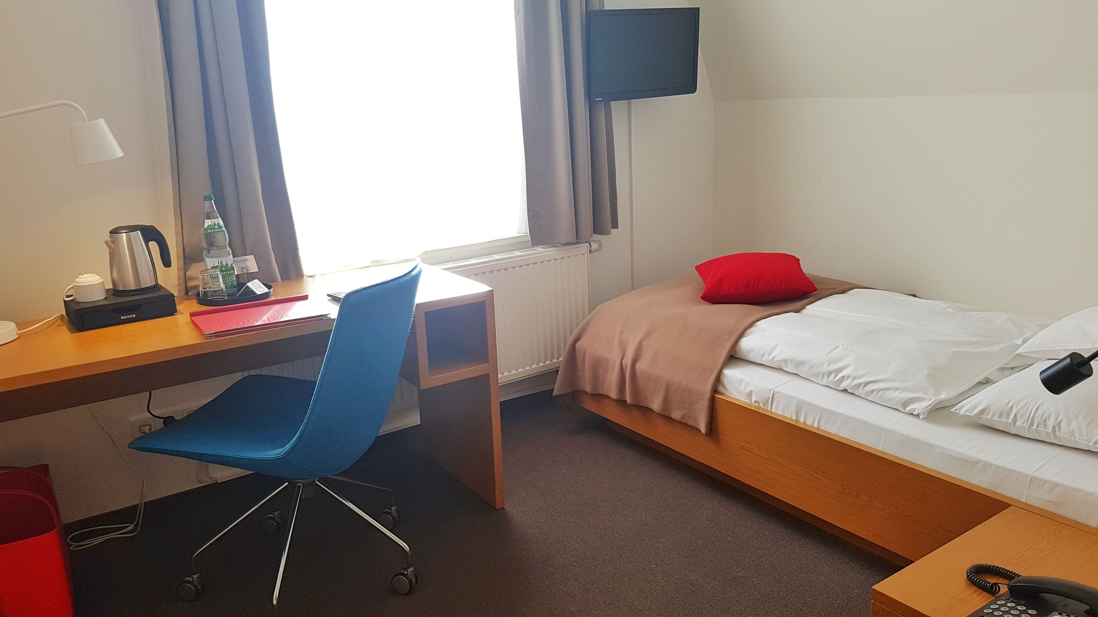 A hotel room with a wooden desk, a blue office chair, a bed with white bedding and a red pillow, a bedside table with a phone, and a window with curtains.