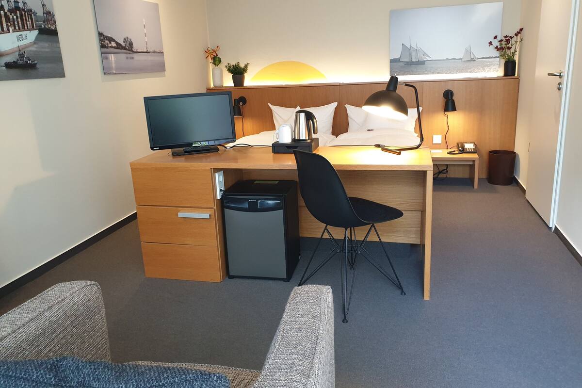 Une chambre d’hôtel avec un bureau en bois, une chaise noire, une télévision, un lit avec une literie blanche et une table de chevet avec une lampe.