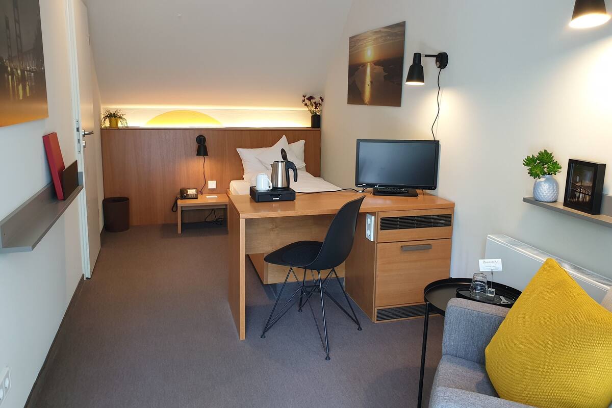 Une chambre d’hôtel moderne avec un bureau en bois, un lit avec une literie blanche, une télévision, une petite table avec une plante et un coussin jaune sur un canapé.