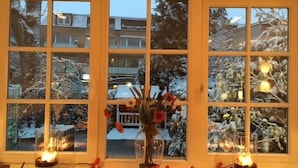 A room with a large window divided into nine panes, each reflecting a snowy outdoor scene. A vase with red flowers is placed on the windowsill, and two candles are lit on the windowsill. - Baurs Park (Hamburg)