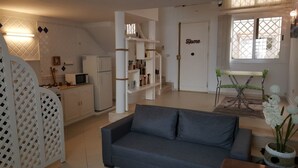 Una sala moderna con un sofá gris, una mesa de comedor con sillas, una zona de cocina con refrigerador y microondas, y una escalera que conduce a otro nivel.