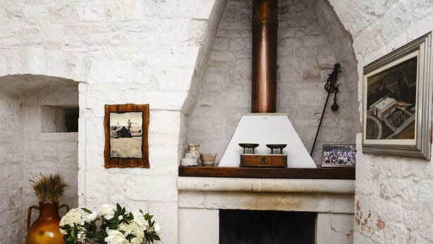 A rustic stone fireplace with a chimney, a framed photograph, and a vase with flowers on a small table. - Masseria Gravelle (Conversano)
