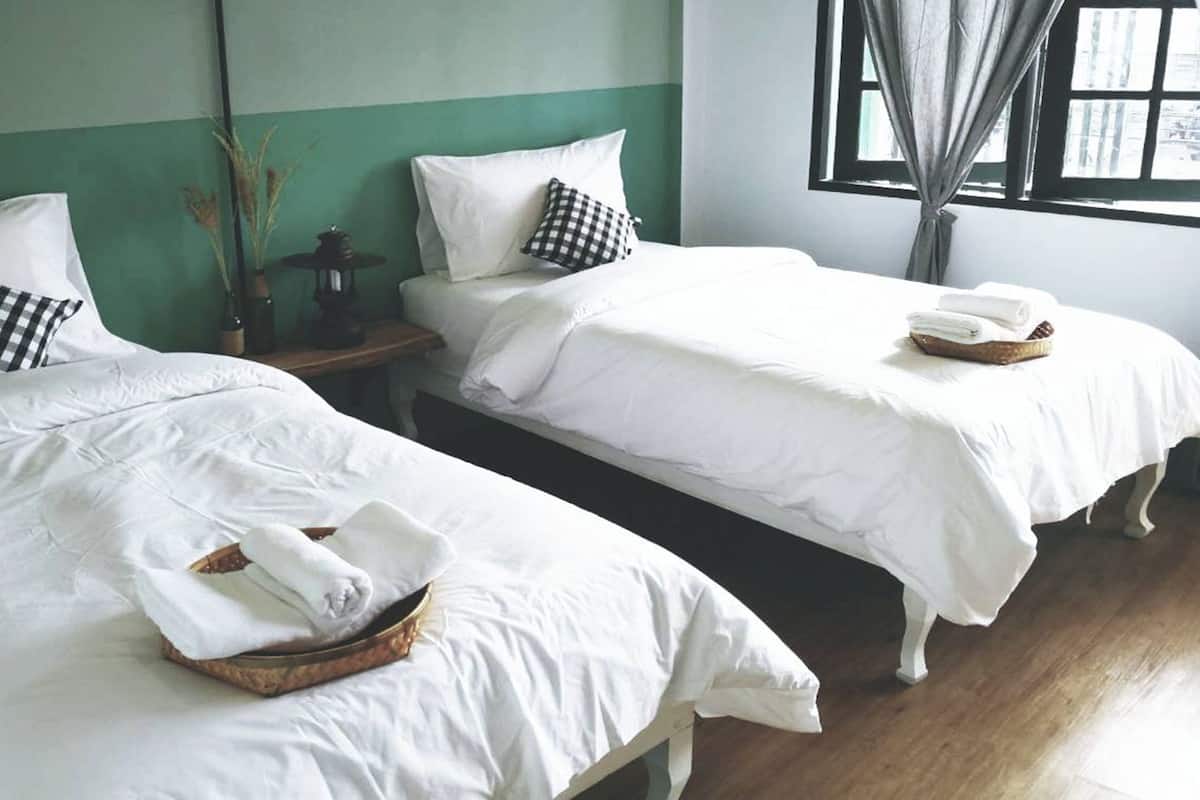 Deux lits simples avec du linge de lit blanc et des oreillers à carreaux, une petite table avec une lanterne et une fenêtre avec des rideaux.