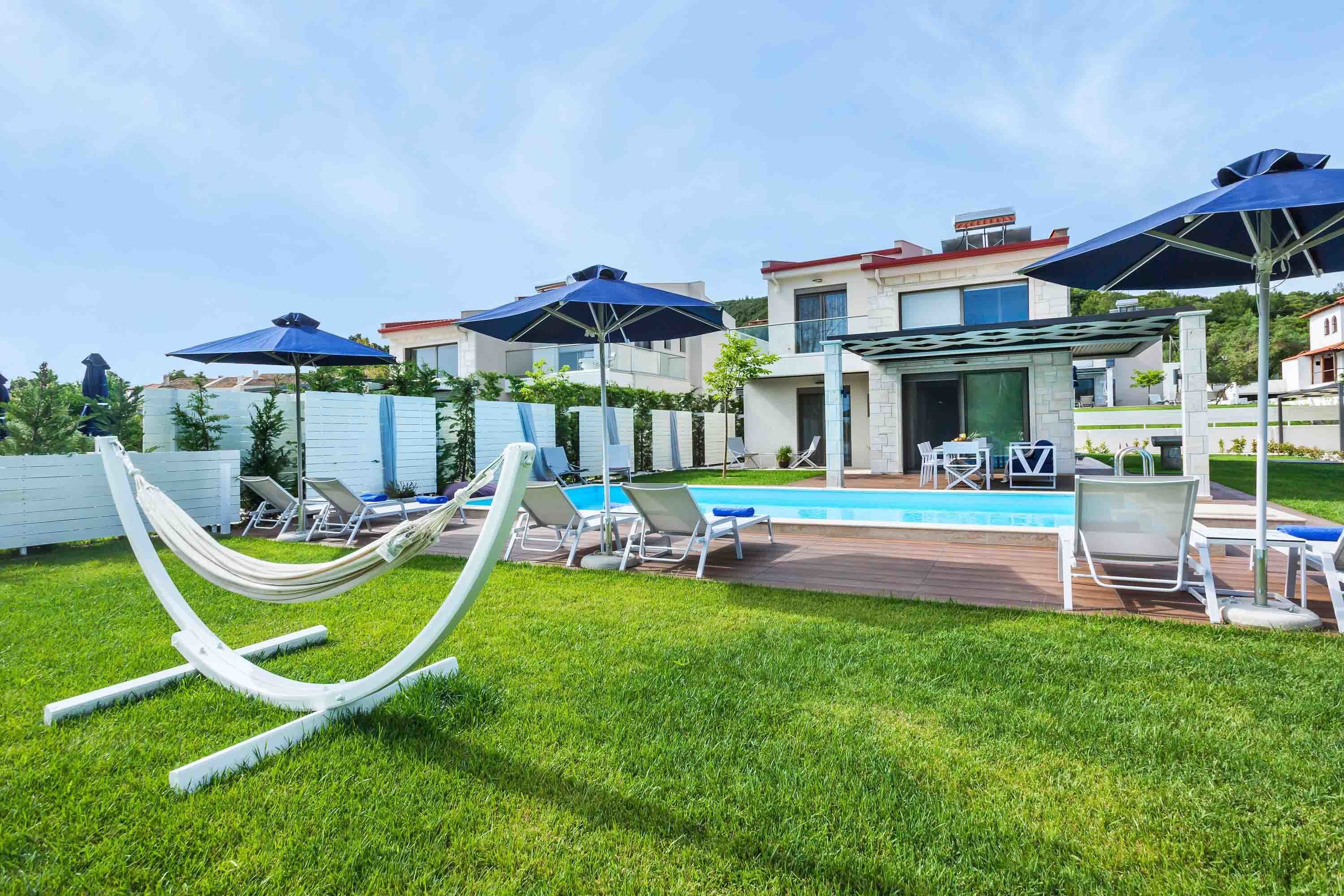 A modern outdoor pool area with lounge chairs and umbrellas, surrounded by well-maintained grass and a poolside deck.