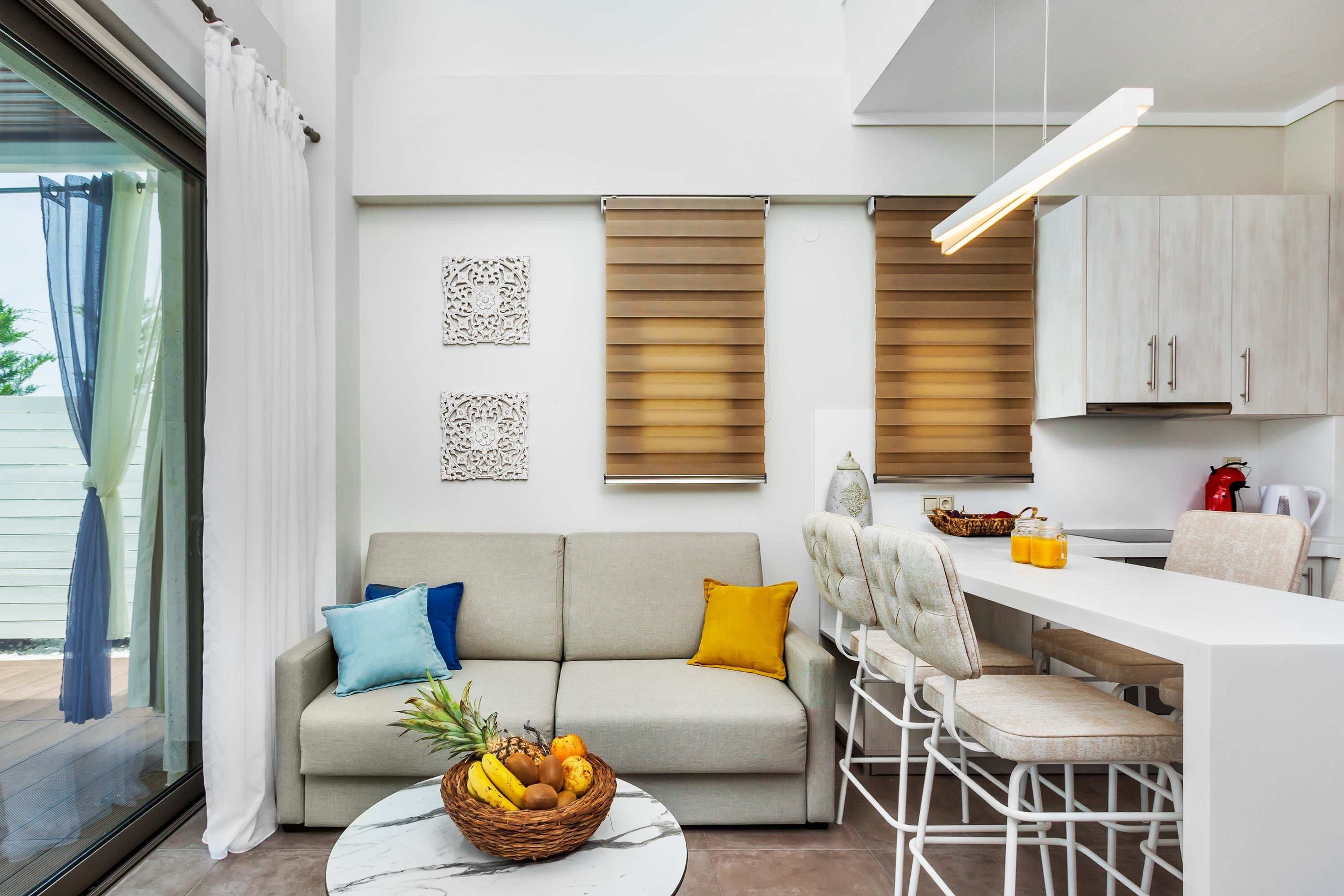 Un salon moderne comprenant un canapé, une table à manger avec des chaises, un panier de fruits et un coin cuisine avec des placards et un luminaire suspendu.
