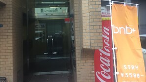 A building entrance with a glass door, a Coca-Cola sign, and a promotional banner for bnb+. -  bnb+ Shinbashi - Hostel (Tokyo)
