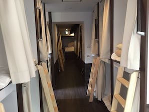 A narrow corridor with bunk beds on either side, numbered from 23 to 24, leading to a room with a bed and a window.