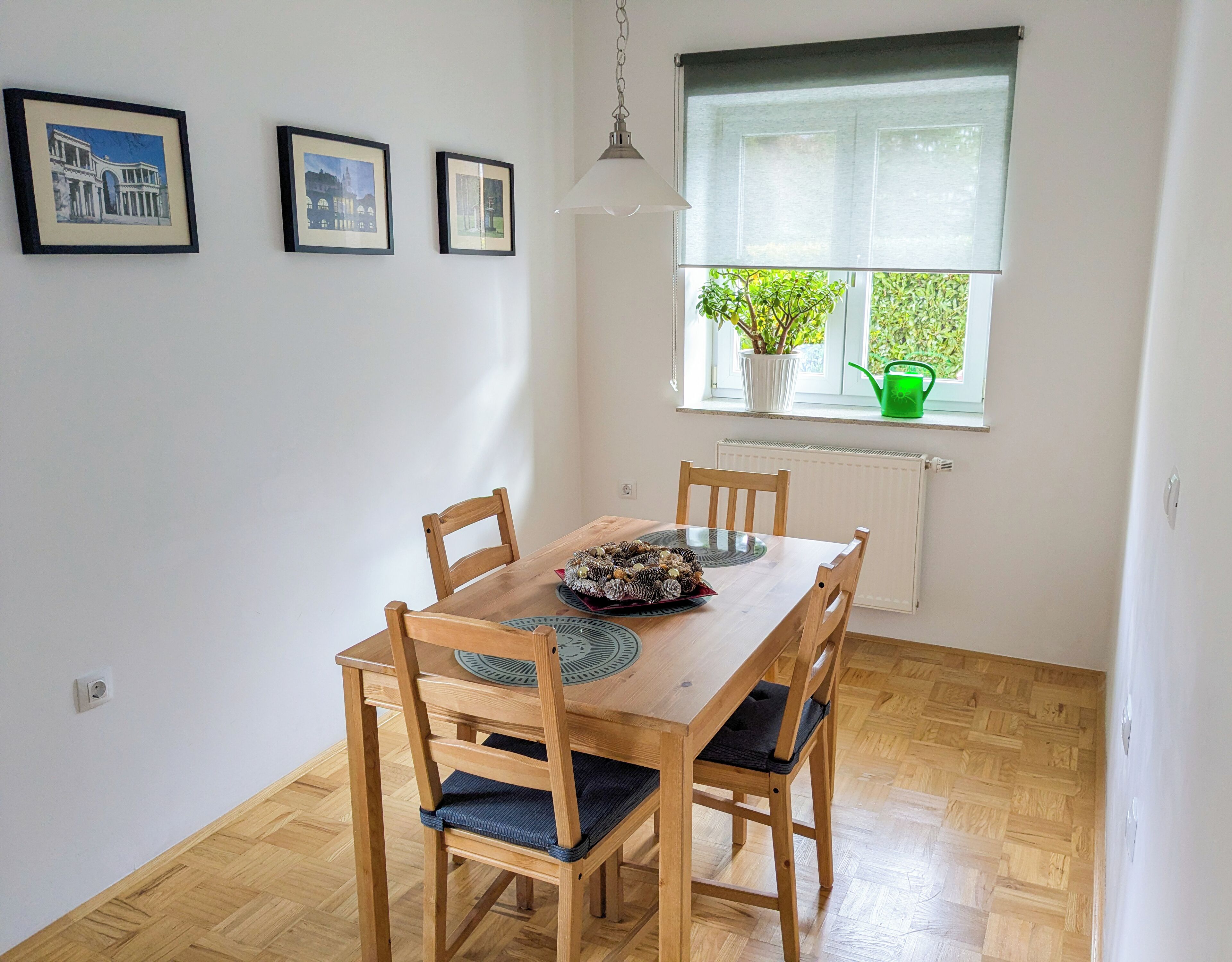 A dining area with a wooden table and chairs, framed pictures on the wall, a window with a view of greenery, and a hanging light fixture.