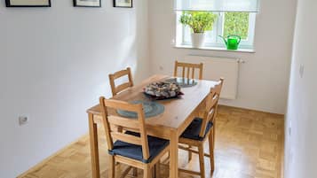 A dining area with a wooden table and chairs, framed pictures on the wall, a window with a view of greenery, and a hanging light fixture.