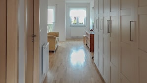 Un pasillo amplio con suelo de madera, paredes blancas y vistas a una sala de estar con sofá y ventana.
