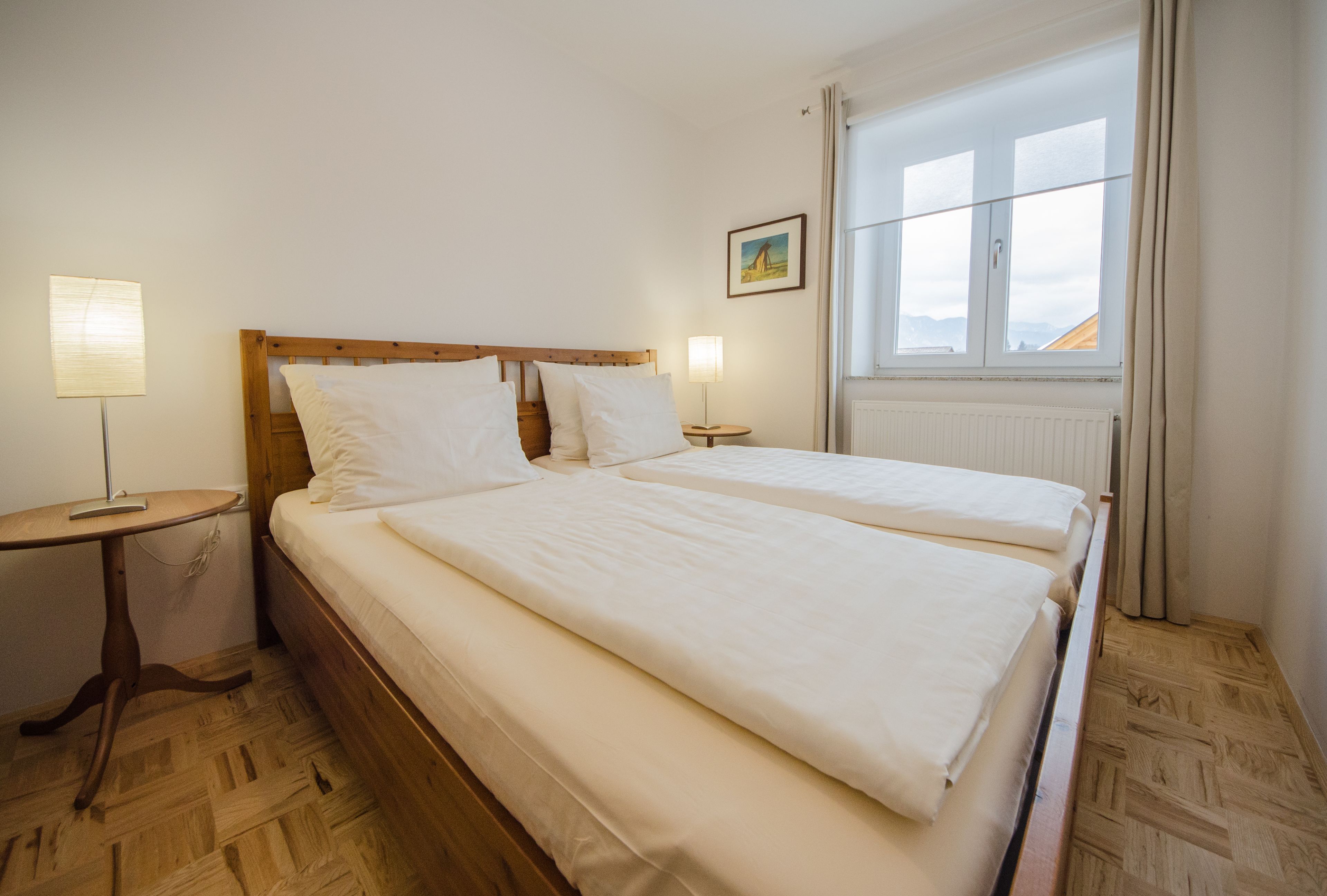 Un llit de matrimoni amb llençols blancs a una habitació amb terra de fusta, una petita taula de nit, una làmpada i una finestra amb vista a les muntanyes.
