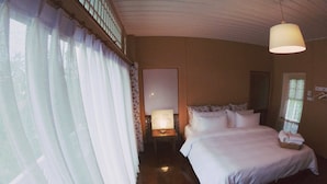 A hotel room with a bed, a bedside table with a lamp, a window with curtains, and a hanging light fixture. - The White Houses (Hua Hin)