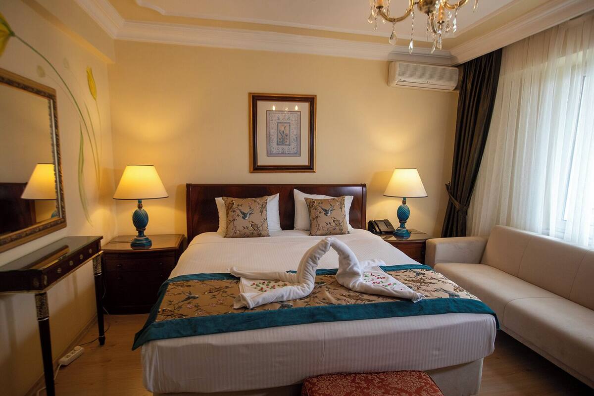 A hotel room with a large bed, two bedside tables with lamps, a sofa, a chandelier, and a framed picture on the wall.
