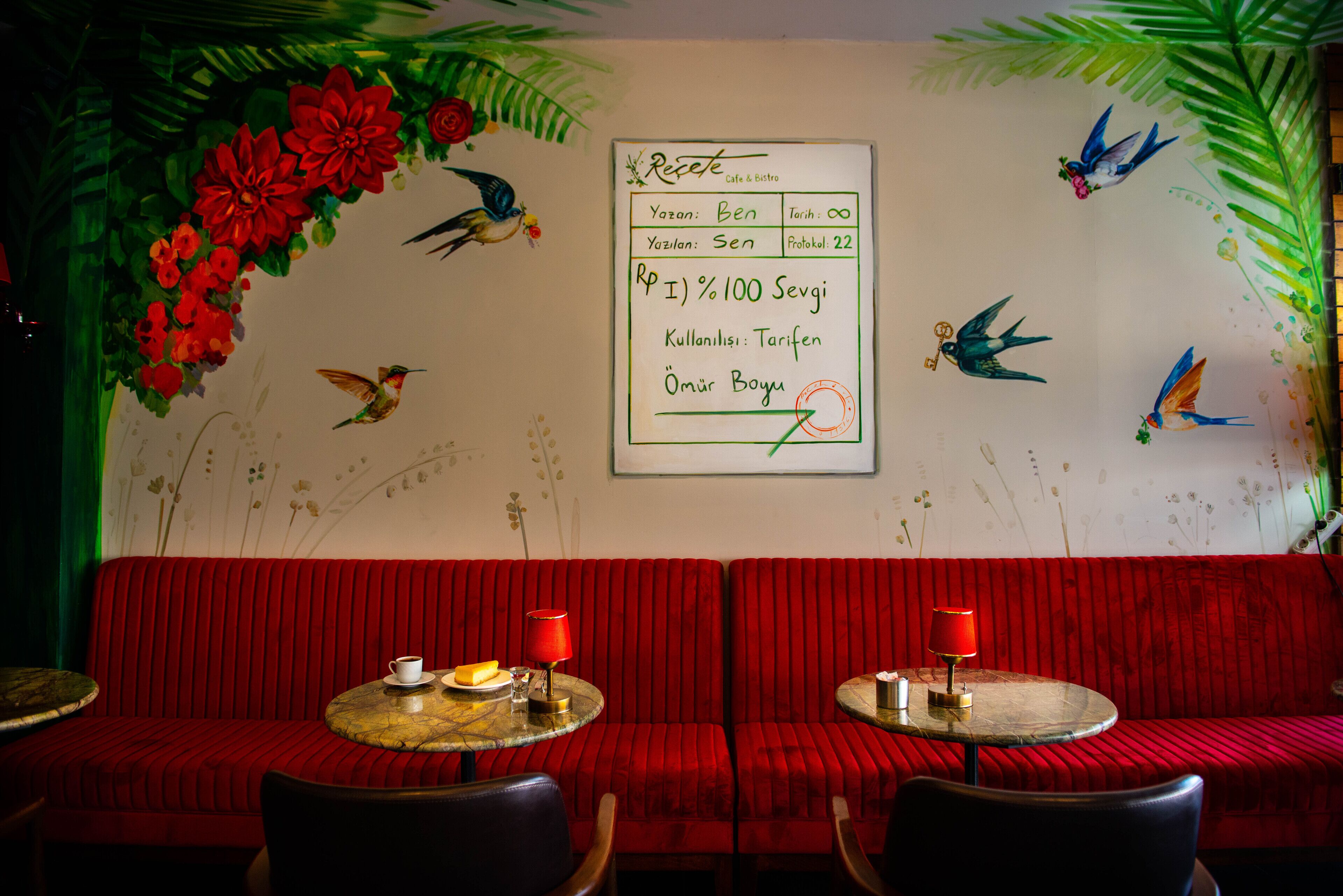 A restaurant interior with red upholstered seating, two round tables with red lamps, and wall decorations including birds and flowers.