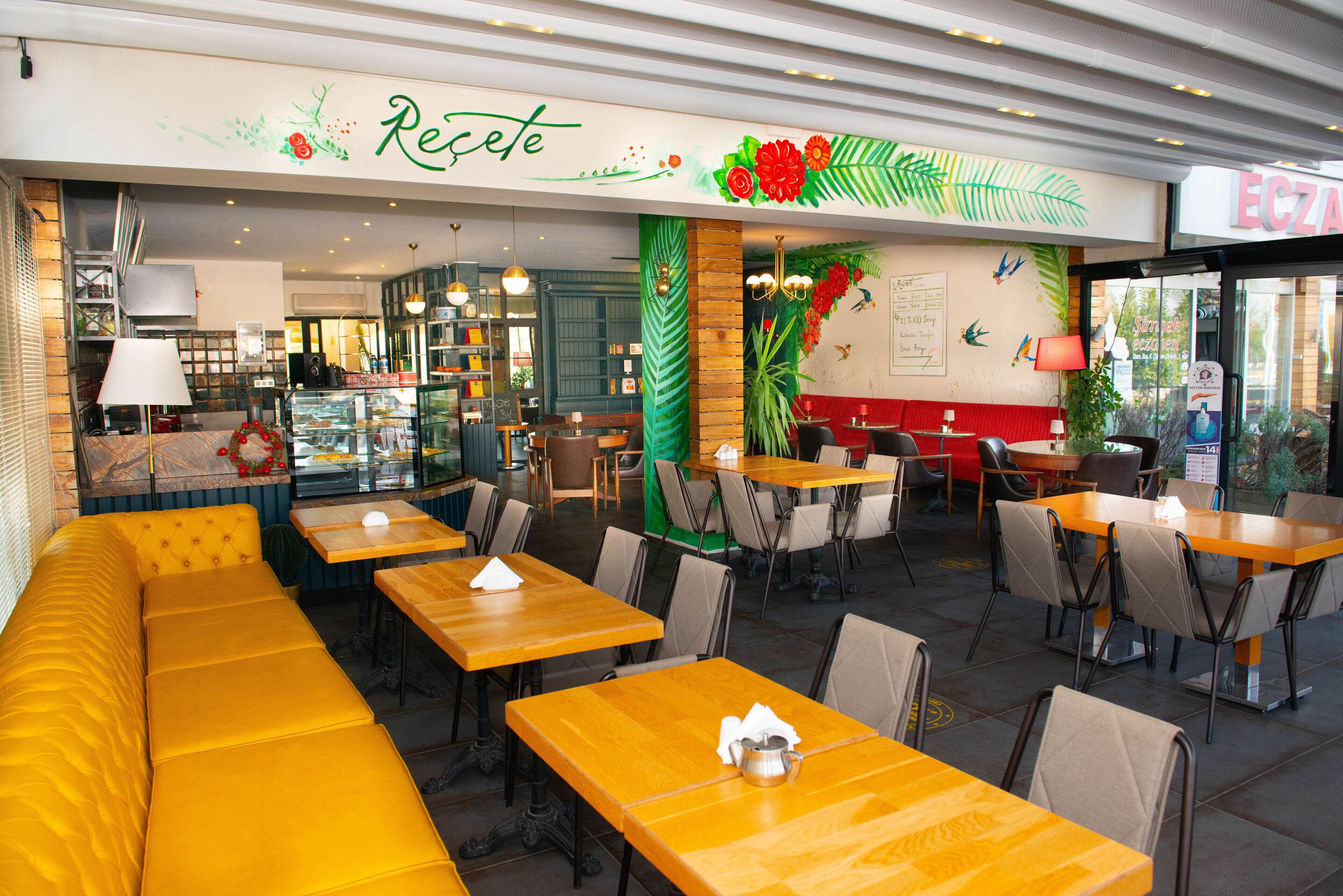 Un restaurante con asientos de color amarillo, mesas de madera y una pared verde con una planta, y un letrero que dice 