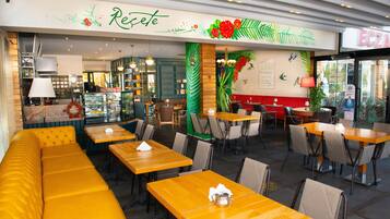 Un restaurante con asientos de color amarillo, mesas de madera y una pared verde con una planta, y un letrero que dice