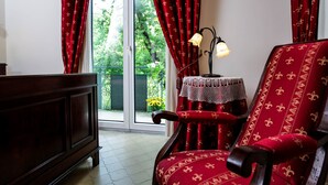 Une pièce avec un meuble en bois, un fauteuil rouge recouvert d’un tissu à motifs floraux, une petite table recouverte d’une nappe en dentelle et une vue sur la verdure à travers une porte vitrée.