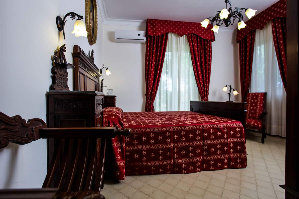 Een slaapkamer met een groot bed, een houten dressoir, een bureau en een stoel. Er is een kroonluchter, een wandlamp en een raam met rode gordijnen.